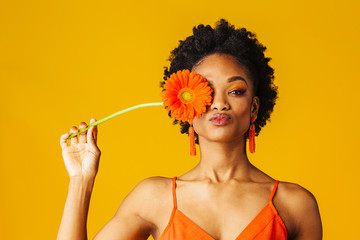Portrait of a y young woman holding orange Gerbera daisy covering her eye and putting lips sending kiss