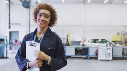 Portrait of female tutor wearing safety glasses teaching auto mechanic apprenticeship at college holding clipboard and smiling at camera - shot in slow motion