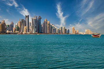 Fototapeta premium Doha Qatar skyline daylight view with clouds in the sky showing skyscrapers and dhow with Qatar flag in background and Arabic gulf in foreground
