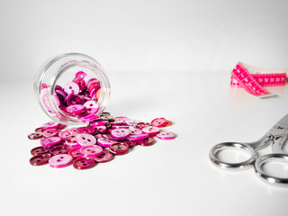 Glass jar full of clothing pink buttons, measuring tape and scissors on white background