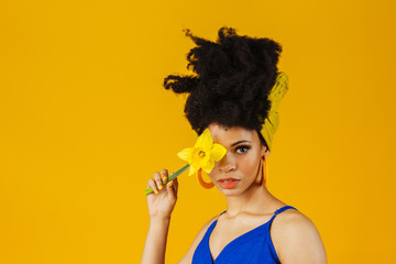 Portrait of young woman in blue holding yellow daffodil flower covering one eye, isolated on yellow...