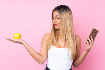 Young Uruguayan woman over isolated pink background having doubts while taking a chocolate tablet in one hand and an apple in the other