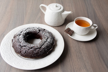 Chocolate cake with a hole in the centre and sugar powder sprinkled on top, on the background are a teapot and teacup with tea