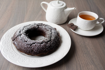Chocolate cake with a hole in the centre and sugar powder sprinkled on top, on the background are a teapot and teacup with tea