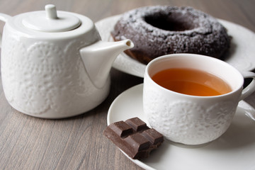 Chocolate cake with a hole in the centre and sugar powder sprinkled on top, on the background are a teapot and teacup with tea