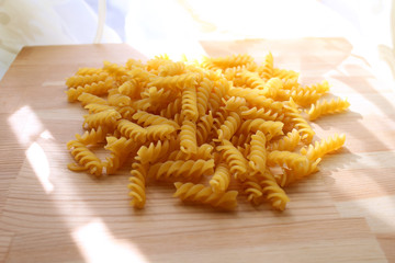 Small mountain of beautiful yellow dry uncooked pasta, poured on light brown wooden surface against background of fabric backlit by sunlight. Backlight, closeup