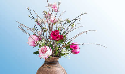 Modern spring flower bouquet with pink and fuchsia Peonies, May bells, Magnolia flowers and willow catkins in an old rustic wooden vase isolated on a blue white background. 