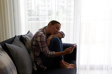 Depressed single woman sitting at home due to worldwide quarantine lockdown. Portrait of lonely female with worried facial expression watching at mobile phone screen. Close up, copy space, background.