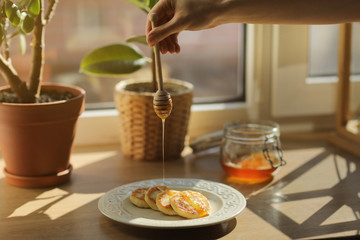 Cheesecakes or syrniki on a plate with honey, in the background a jar with honey and ficus. a woman pouring wooden spoon for honey which flows honey. Close up. selective focus