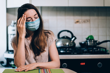 A woman with a cold sits at the table in the kitchen wearing a coronavirus mask and holding her...