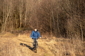 A teenager rides a Bicycle on dry grass against a background of bare trees. Early spring landscape.