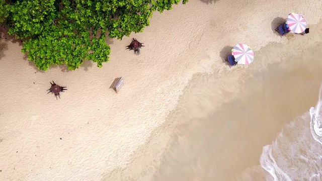 Aerial still footage of white-sand beach with waves coming in and out in the corner. Two umbrellas on the beach and tree leaves. 