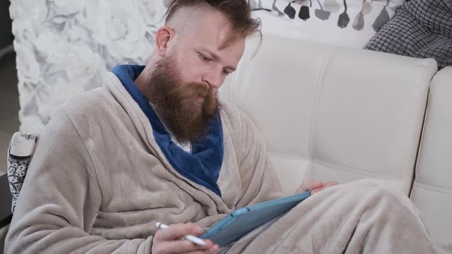 man in housecoat with beard and mustache is lying on sofa and drawing on tablet question mark,working at home,designer and artist in quarantine of Coronavirus global epidemic.4 K