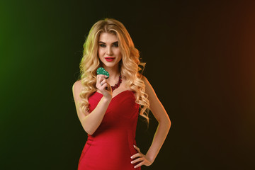 Blonde girl in red fitting dress and necklace. She smiling, showing two green chips, posing on colorful background. Poker, casino. Close-up