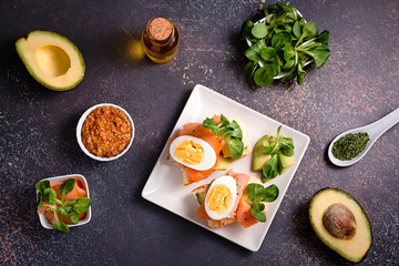 Healthy toasts with smoked salmon and avocado served with mustard, eggs and leafy greens on black table top. Mediterranean diet snack, open sandwich