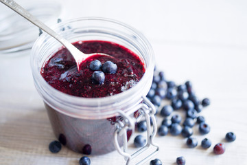 Fresh bilberry and blueberry jam in a glass jar on a white wooden surface, close-up