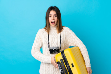 Traveler woman holding a suitcase over isolated blue wall with surprise and shocked facial...