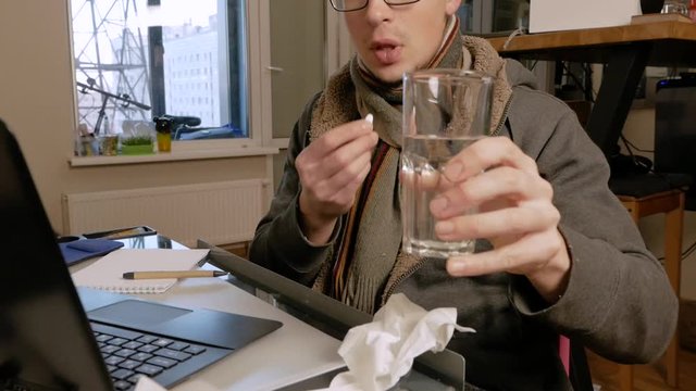 A young man in glasses takes a tablet, drinks it with water and works on a laptop at home.