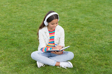write song lyrics. happy childhood concept. kid write memory in diary. schoolgirl relax with notebook. happy child listen music in headphones. small girl on green grass. kid enjoy song in park nature