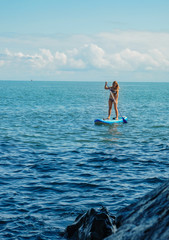 SUP Stand up paddle board. Blond girl stand with paddle in the sea near stone beach. Blue sea with waves Mtsvane Kontskhi Beach, Batumi, Georgia.