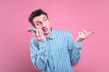 Picture of skunny slim young man in blue shirt talking. Conversation wireless through smartphone. Isolated over pink background.