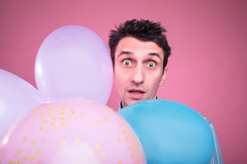 Amazed confused young man look straight and pose with colorful balloons in front. Isolated over pink background.