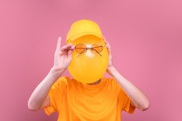Funny picture of incognito guy hiding face behind yellow balloon. Hold sunglasses in front of it. Guy wear yellow cap and shirt. Isolated over pink background.