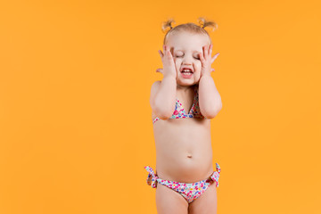 a girl in a bright swimsuit clenches her cheeks with her hands, making funny faces and closing her...