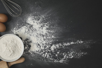 Wooden dough roller, a cup of flour and whisk on the black table with flour spread.