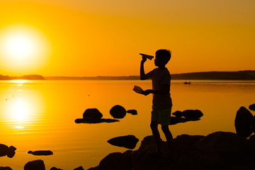 Fototapeta na wymiar Little boy holds a paper plane in hand at sunset. A child raised his hand up to sky and plays with origami in the evening at lake. Silhouette