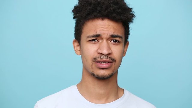 Unshaven african american guy 20s in white t-shirt curly hair isolated on blue background studio portrait. People lifestyle concept. Look at camera say hush be quiet with finger on lips shhh gesture