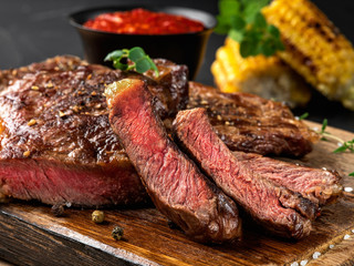 Sliced, fried, spiced steaks with herbs on wooden board, grilled corn, red sauce in small dark bowl on a black background. Close-up shot. Side view.