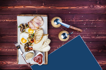 Big breakfast. Ham, bread toast, fried eggs, grilled mushrooms and tomatoes with sauce. There is a pepper grinder, cutlery and a place for text. Top view