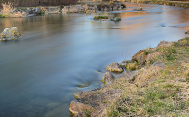 A river flows quietly and slowly in the winter between dry grass and stones as a long-term shot