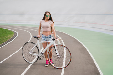 Naklejka premium Cycling and fitness. Young beautiful fit woman with perfect body in sportswear with the bike outdoors on the track. Sportive and healthy lifestyle, cyclist working out, training, fashion, beauty.