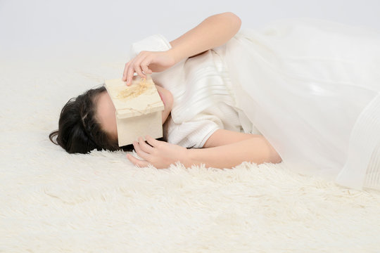 muchacha con vestido blanco acostada sobre el suelo y con una libreta ocultándose la cara
