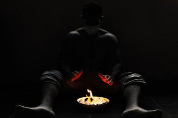 Hombre sentado en el suelo con mascarilla calentándose las manos con fuego