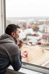 A young man in a medical mask looks out of the window of his house at the panorama of the city. A dark-haired man looks out of a window at an empty city during a coronavirus epidemic