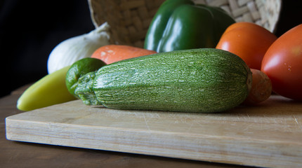 close up or macro photography of a zucchini ,squash with assorted vegeatbles on the back, tomatoes, onion, chile, pepper and a basket in the background, dark food.