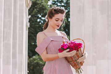Sensual attractive young vintage woman in pink stripped dress and pinup hairstyle holding basket of peonies flowers in hands outdoors. Beautiful stylish retro girl walking in summer park. Retro outfit