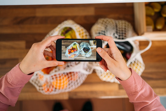 Top view of a woman taking photos with the smartphone of fruits and vegetables in a reusable organic cotton mesh bag. Zero waste, plastic free concept. Sustainable lifestyle.