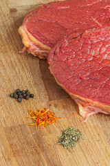 Slices of a raw fresh meat are on the wooden desktop with a peppercorns, saffron and rosemary spacies. Concept of healthy dinner. Ingredients for cooking beef steaks.