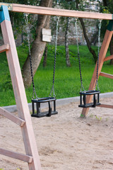 A swing consisting of two chairs for small children with a safety system, suspended from metal chains and sand on the ground on the Playground against a background of green grass. Swing without childr