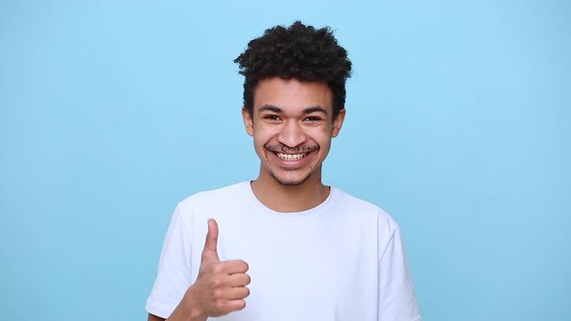 Unshaven smiling african american young guy 20s in white t-shirt curly hair posing isolated on blue background studio portrait. People lifestyle concept. Looking at camera showing thumbs up gesture