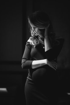 Beautiful young sexy pregnant woman with curls and make-up in a black tight-fitting dress. Black and white photo. Fine art. Waiting for a child. Soft selective focus.