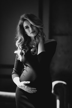 Beautiful young sexy pregnant woman with curls and make-up in a black tight-fitting dress. Black and white photo. Fine art. Waiting for a child. Soft selective focus.