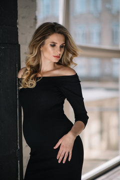 Beautiful young sexy pregnant woman with curls and make-up in a black tight-fitting dress. Expectation of a child of 9 months. Soft selective focus.