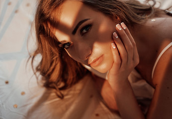 Close-up portrait of a confident and sexy female model with brown curly hair wearing white lingerie while laying on bed among sheets and pillows, looking smooth