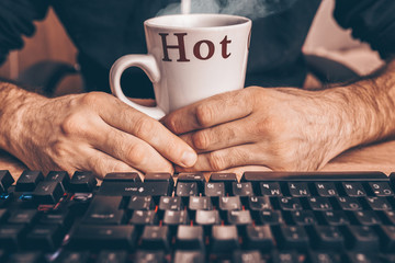A cup of hot drink with the inscription Hot in hands near a computer - remote work and study