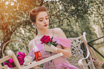 Portrait of young tender beautiful woman with pin up hairstyle and pink vintage dress having picnic on summer garden. Attractive gorgeous stylish retro girl drink posing with peonies flowers outdoors
